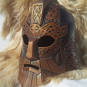 Peut inclure: Un casque détaillé, sculpté avec soin, avec une barbe et des motifs complexes. Le casque est fait de matériaux brun foncé et dorés.