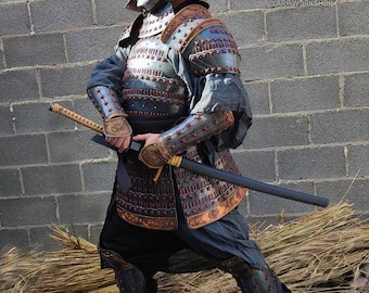 May include: A full suit of samurai armor featuring a metallic helmet with a crest, a chest plate with red lacing, shoulder guards, arm bracers, and leg guards. The armor is worn over dark clothing and includes a long sword with a black scabbard.
