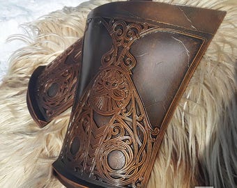 May include: A pair of brown leather arm guards with intricate gold-colored designs. The guards have a textured surface and are decorated with swirling patterns.