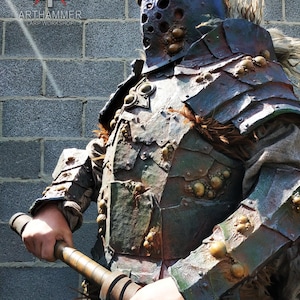 Puede incluir: Una figura detallada, fuertemente armada, con un casco que presenta una máscara facial con agujeros y una gran pieza de pecho verde y marrón tachonada. La figura sostiene un gran garrote de madera.