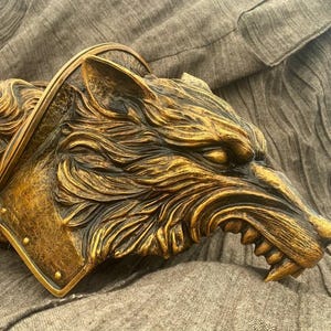 May include: A golden wolf head sculpture with a detailed, textured surface. The wolf's mouth is open, revealing sharp teeth. The sculpture is mounted on a textured, brown surface.