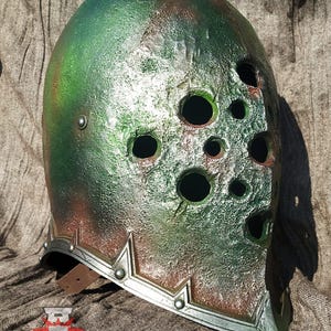 May include: A green and brown helmet with a silver trim and multiple holes. The helmet has a leather strap on the side.
