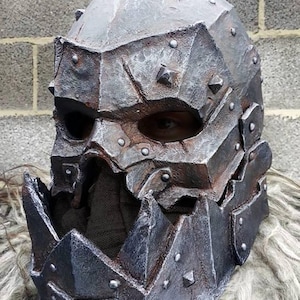 Puede incluir: Un casco de metal detallado, hecho a mano, con un acabado oxidado y desgastado. El casco tiene una parte superior puntiaguda y una máscara facial con una sola abertura para el ojo. El casco está hecho de metal y tiene una superficie texturizada. El casco es un accesorio para LARP o cosplay.