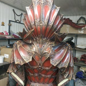 Puede incluir: Una armadura roja y dorada detallada y adornada con un gran casco elaborado. La armadura está hecha de metal y tiene un diseño texturizado e intrincado.