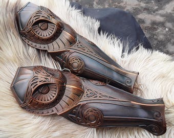 May include: Pair of bronze-colored arm guards with intricate, ornate designs. The armor features detailed patterns and a weathered finish, suggesting a historical or fantasy theme. The arm guards are displayed on a fluffy, light-colored surface.