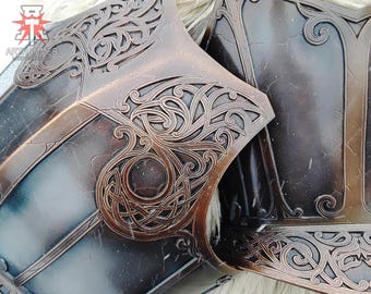May include: Close-up of a decorative metal armor piece with intricate, ornate designs. The armor is a dark bronze color with a textured surface. The design features a repeating pattern of swirls and knots.