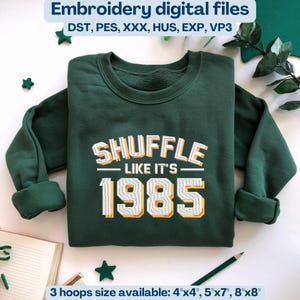 Op de afbeelding: Een donkergroene sweatshirt met de geborduurde tekst "SHUFFLE LIKE IT'S 1985" in wit en geel. De afbeelding bevat ook de tekst "Embroidery digital files" en "3 maten borduurringen beschikbaar: 10cm x 10cm, 13cm x 18cm, 20cm x 20cm".