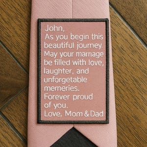 May include: A pink tie with a black embroidered patch that reads "John, As you begin this beautiful journey. May your marriage be filled with love, laughter, and unforgettable memories. Forever proud of you. Love, Mom & Dad"
