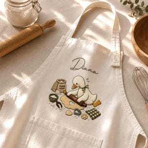 May include: A cream-colored cotton apron featuring an embroidered illustration of a white duck baking with a rolling pin, flour bag, and heart-shaped cookies. The name Diana is embroidered in dark green script above the duck graphic.