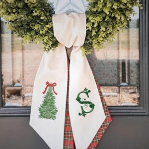 Puede incluir: Un colgador de puerta de tela color crema con un árbol de Navidad bordado en verde con un lazo rojo a la izquierda y una letra S bordada en verde a la derecha, con un reverso de tela de cuadros rojos y verdes visible en los bordes.