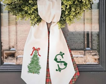May include: A cream-colored fabric door hanger featuring a green embroidered Christmas tree with a red bow on the left side and a green embroidered letter S on the right side, with a red and green plaid fabric backing visible at the edges.