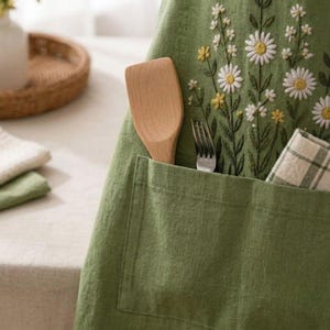 May include: An olive green apron featuring embroidered white and yellow daisy flowers. The apron has a large front pocket containing a wooden spatula, a metal fork, and a folded white and green checkered cloth napkin for kitchen use.