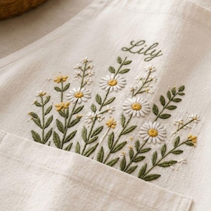 May include: An off-white apron featuring detailed embroidery of white daisies with yellow centers and green leaves. The name Lily is embroidered in cursive green script above the floral arrangement, which extends from the top of a front pocket.