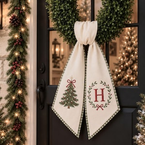 Peut inclure: Un porte-porte en tissu crème avec un sapin de Noël brodé en vert et rouge d'un côté et une lettre H rouge dans une couronne verte et rouge de l'autre. Le tissu présente une bordure à carreaux verts et blancs.