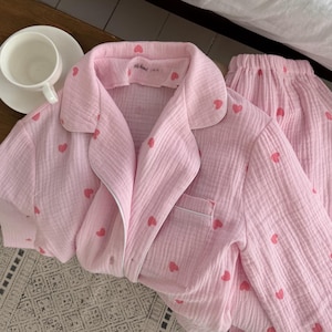 Puede incluir: Conjunto de pijama rosa con estampado de corazones rojos. El conjunto incluye una camisa con cuello y botones con bolsillo en el pecho y pantalones a juego. Una taza y un platillo blancos están sobre una superficie de madera.