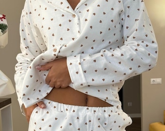 May include: A white long-sleeve pajama set featuring a repeating pattern of small brown hearts. The top has a collared neckline with a button-down front, and the matching pants feature an elasticized waistband for a comfortable fit.
