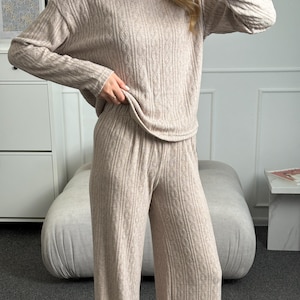 Op de afbeelding: Een beige tweedelige loungeset bestaande uit een top met lange mouwen en een broek met wijde pijpen. Beide kledingstukken hebben een verticale kabelgebreide textuur, een relaxte pasvorm, een ronde halslijn en een elastische tailleband.