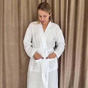 May include: A white waffle-weave robe with a sash tie and two pockets. The robe is worn by a person standing in front of a brown curtain.