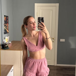 May include: A two-piece set in a dusty rose color, featuring a V-neck crop top and ruffled shorts. The fabric has a textured, crinkled appearance. The outfit is designed for comfort and casual wear.