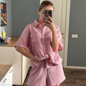 Op de afbeelding: Een lichtroze tweedelige pyjamaset. De set bestaat uit een overhemd met korte mouwen en knopen met een borstzak en bijpassende shorts. De stof heeft een getextureerde, gekreukelde uitstraling.