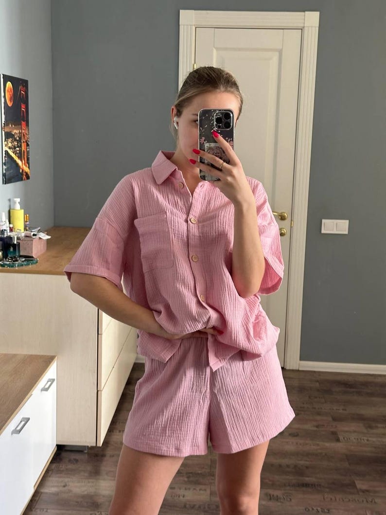 Op de afbeelding: Een lichtroze tweedelige pyjamaset. De set bestaat uit een overhemd met korte mouwen en knopen met een borstzak en bijpassende shorts. De stof heeft een getextureerde, gekreukelde uitstraling.