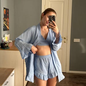 May include: A light blue pajama set consisting of a robe, a bralette, and ruffled shorts. The robe has wide sleeves and a draped front. The shorts have a ruffled hem. The set is made of a lightweight, textured fabric.