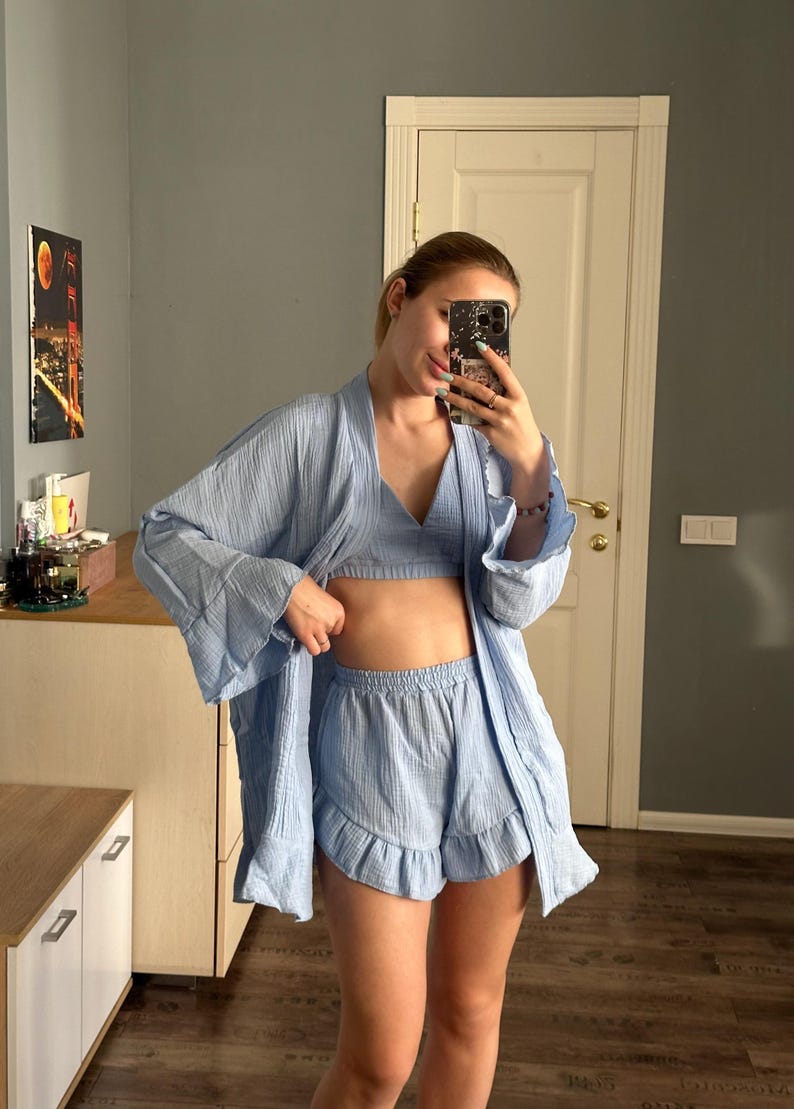 May include: A light blue pajama set consisting of a robe, a bralette, and ruffled shorts. The robe has wide sleeves and a draped front. The shorts have a ruffled hem. The set is made of a lightweight, textured fabric.