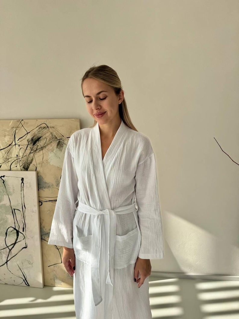 May include: A white, textured robe with a wrap-around design and a matching belt. The robe has long sleeves and two front pockets. The fabric appears lightweight and comfortable. The robe is suitable for lounging or spa use.