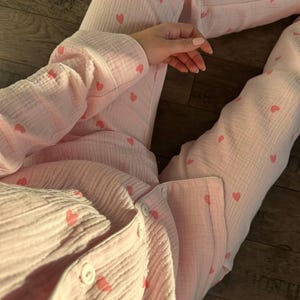 Puede incluir: Conjunto de pijama rosa claro con estampado de corazones. El conjunto incluye una camisa con botones y pantalones. La tela tiene un aspecto texturizado. Los pijamas están salpicados de pequeños corazones rosas.