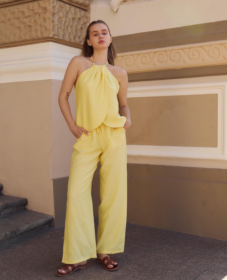May include: A light yellow halter top and wide-leg pants set. The top has a gathered neckline and the pants have a relaxed fit. The outfit is made of a textured fabric. Brown sandals complete the look.