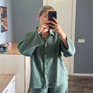 Op de afbeelding: Een tweedelige saliegroene pyjamaset. De blouse met lange mouwen en knopen heeft een borstzak en witte knopen. De bijpassende broek heeft wijde pijpen. De stof lijkt een getextureerde katoen- of linnenmix te zijn. De set wordt in een goed verlichte kamer gepresenteerd.