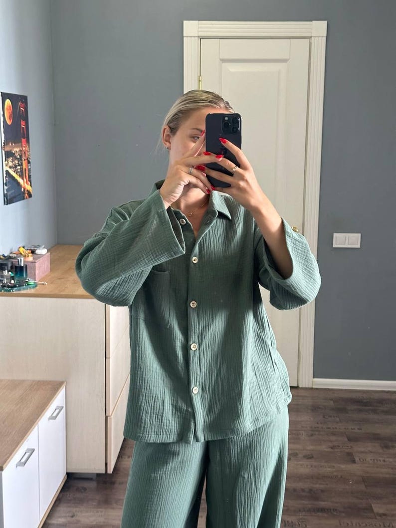 Op de afbeelding: Een tweedelige saliegroene pyjamaset. De blouse met lange mouwen en knopen heeft een borstzak en witte knopen. De bijpassende broek heeft wijde pijpen. De stof lijkt een getextureerde katoen- of linnenmix te zijn. De set wordt in een goed verlichte kamer gepresenteerd.