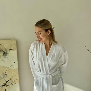 May include: A white, textured robe with a V-neck, long sleeves, and a matching belt. The robe has two front pockets and is made of a lightweight fabric. The robe is designed for comfort and relaxation.