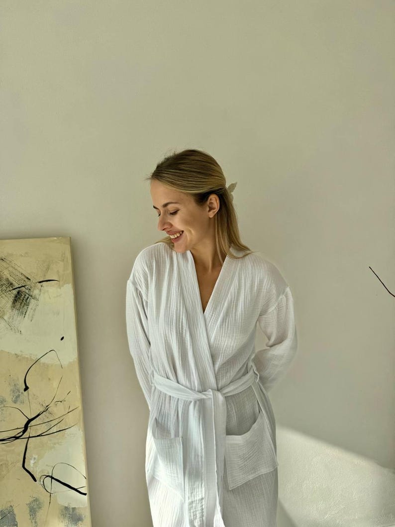 May include: A white, textured robe with a V-neck, long sleeves, and a matching belt. The robe has two front pockets and is made of a lightweight fabric. The robe is designed for comfort and relaxation.