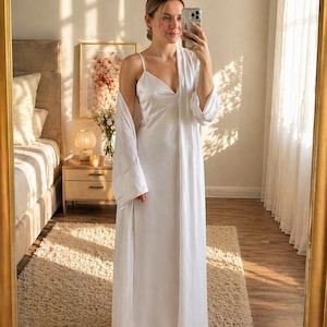 May include: A white satin slip dress with spaghetti straps and a matching long robe. The robe has wide sleeves. The woman is standing in front of a mirror. The bedroom has a neutral colour palette with a beige rug.