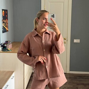 Op de afbeelding: Een stofroze pyjamaset, bestaande uit een overhemd met knopen en een borstzak en bijpassende shorts. De stof lijkt een getextureerde katoen- of linnenmix te zijn. De set wordt door een persoon in een selfie gedragen.