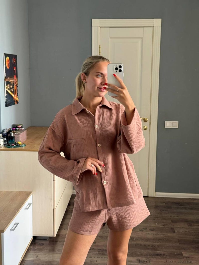 Op de afbeelding: Een stofroze pyjamaset, bestaande uit een overhemd met knopen en een borstzak en bijpassende shorts. De stof lijkt een getextureerde katoen- of linnenmix te zijn. De set wordt door een persoon in een selfie gedragen.