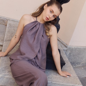 May include: A mauve halter top and matching pants. The top has a draped neckline and the pants are wide-legged. The outfit is made of a textured fabric. The model is sitting on a stone ledge next to a black lamp post.