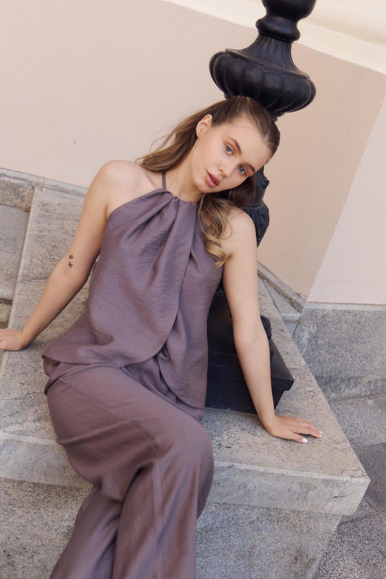 May include: A mauve halter top and matching pants. The top has a draped neckline and the pants are wide-legged. The outfit is made of a textured fabric. The model is sitting on a stone ledge next to a black lamp post.