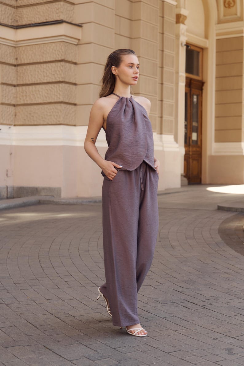 May include: A two-piece mauve outfit featuring a halter top and wide-leg pants. The top has a gathered neckline, and the pants have a drawstring waist. The outfit is made of a textured fabric. The model is wearing white high-heeled sandals.