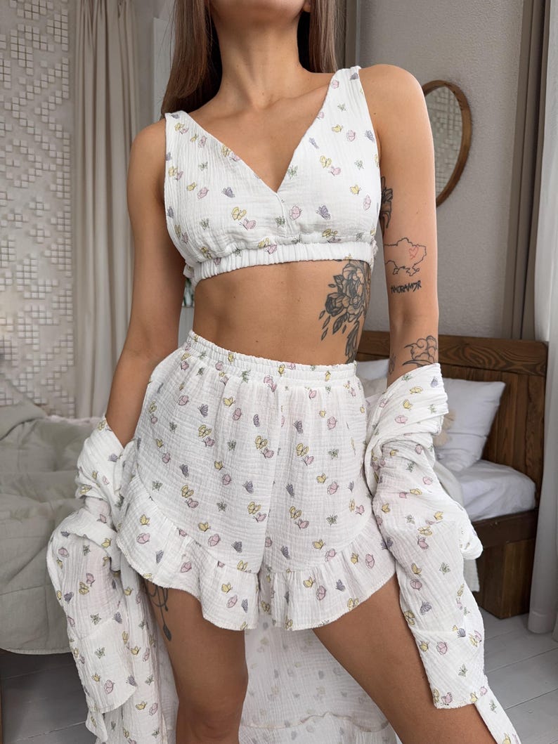 May include: White pajama set with a butterfly print. The set includes a V-neck crop top, ruffled shorts, and a long-sleeved shirt. The fabric appears to be a lightweight, textured material. The butterflies are in pastel colors.
