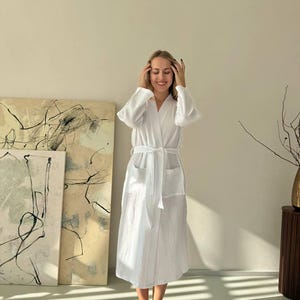 May include: A white, textured robe with a matching belt and pockets. The robe is knee-length and has long sleeves. The person is wearing white fluffy slippers. The background includes abstract art and a wooden side table.