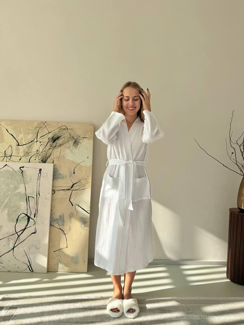 May include: A white, textured robe with a matching belt and pockets. The robe is knee-length and has long sleeves. The person is wearing white fluffy slippers. The background includes abstract art and a wooden side table.