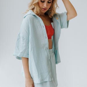 Op de afbeelding: Een lichtblauwe tweedelige outfit bestaande uit een shirt met korte mouwen en bijpassende shorts. Het shirt is open en onthult een rode beha. De outfit is gemaakt van een textuurstof.