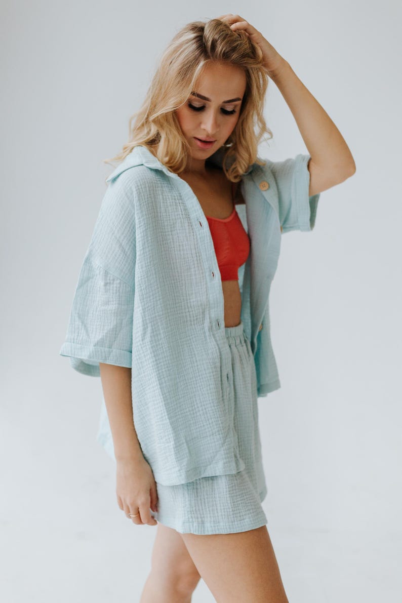 Op de afbeelding: Een lichtblauwe tweedelige outfit bestaande uit een shirt met korte mouwen en bijpassende shorts. Het shirt is open en onthult een rode beha. De outfit is gemaakt van een textuurstof.