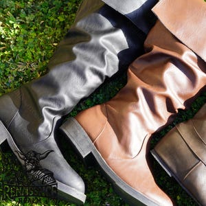 May include: Two pairs of knee-high boots, one black and one brown, are laid out on a bed of green grass. Both boots have a slouchy, relaxed fit and are made of a faux leather material.