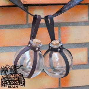May include: Two clear glass bottles with cork stoppers and brown leather straps hanging from a black leather strap. The bottles are round and have a rustic look.