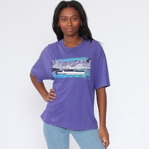 May include: A purple t-shirt with a graphic of a boat and the text "Niagara Falls" on the front. The model is wearing light blue jeans and white sneakers.