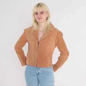 May include: A woman wearing a light brown suede jacket with a zipper closure and light blue jeans. She is wearing white sneakers.