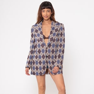 May include: A long-sleeved blazer with a diamond pattern in shades of blue, brown, and beige. The blazer has a notched collar and two front pockets. The model is wearing white trainers.